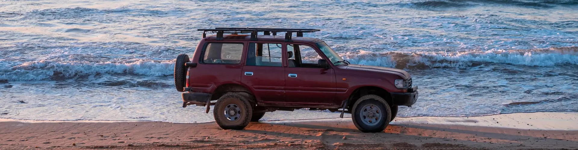 A dark red four-wheel-drive parked on wet sand at the edge of the surf