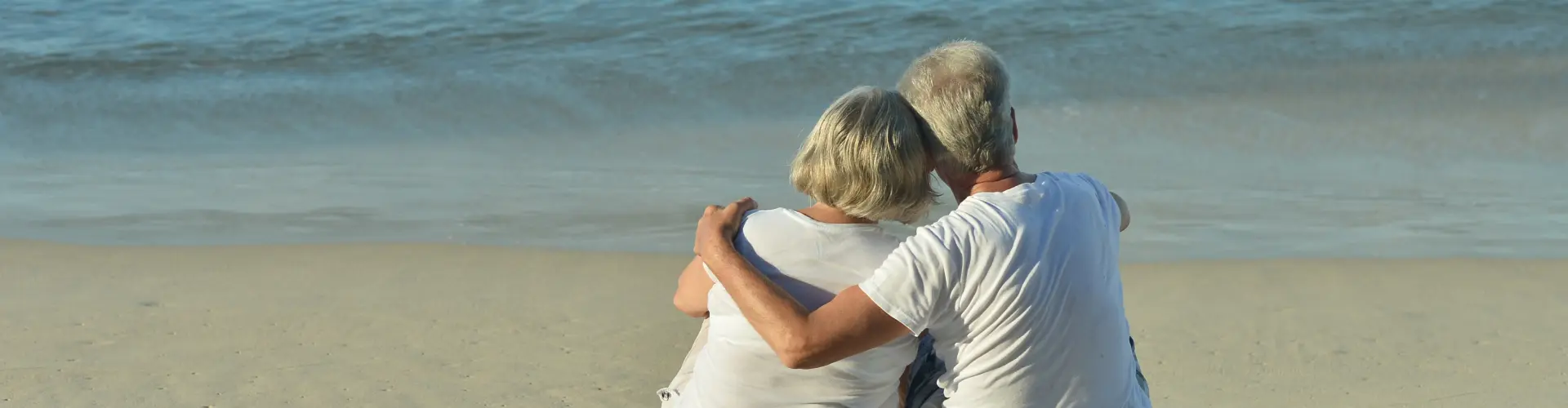 An older couple sitting close on the sand, arms around one another, looking out at a calm sea