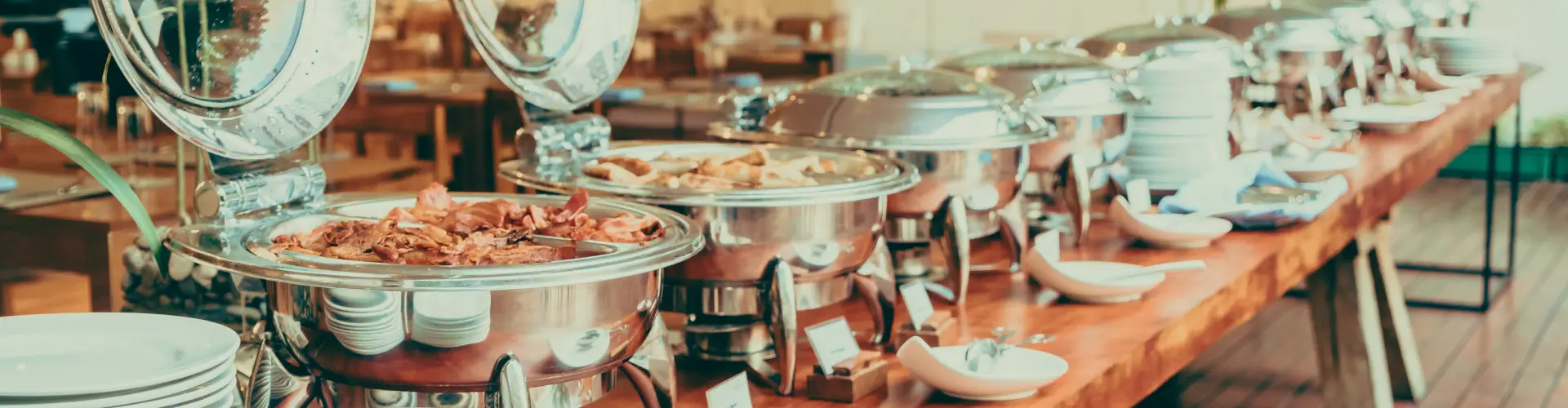 A buffet counter of silver chafing dishes with bacon and pastries, stacked white plates along a wooden table