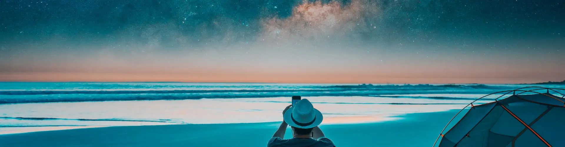 A man in a straw hat photographing the Milky Way over the sea, a dome tent pitched on the sand beside him