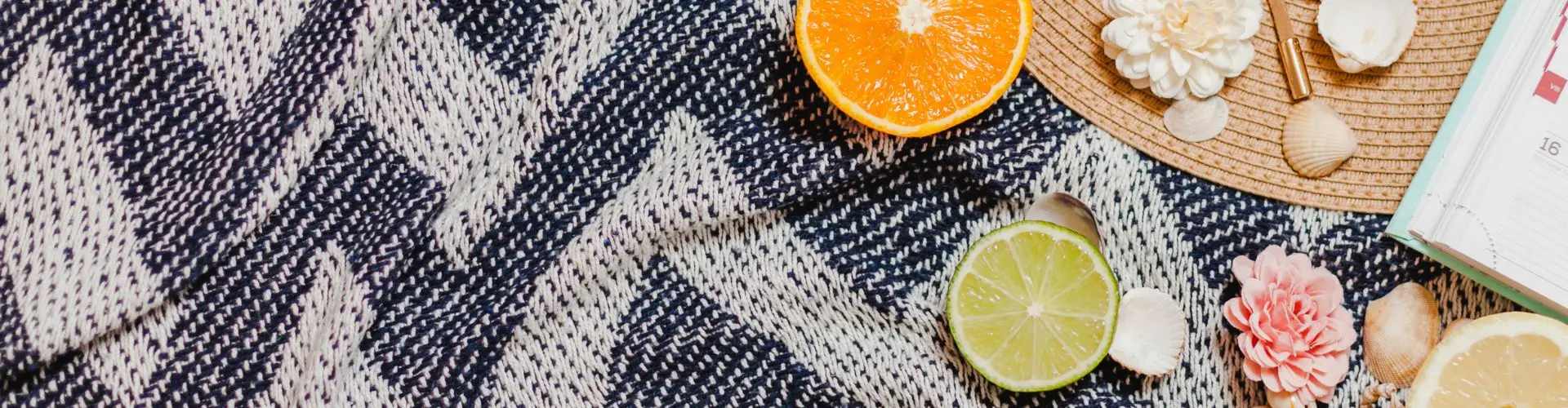 A flat lay on a blue chevron blanket of orange and lime slices, seashells and a pink bloom on a woven mat