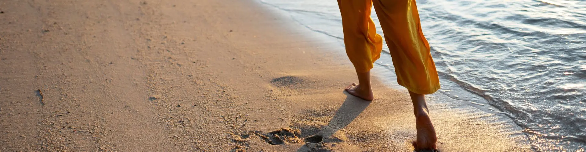 Bare feet below orange trousers walking along the wet sand at the water's edge