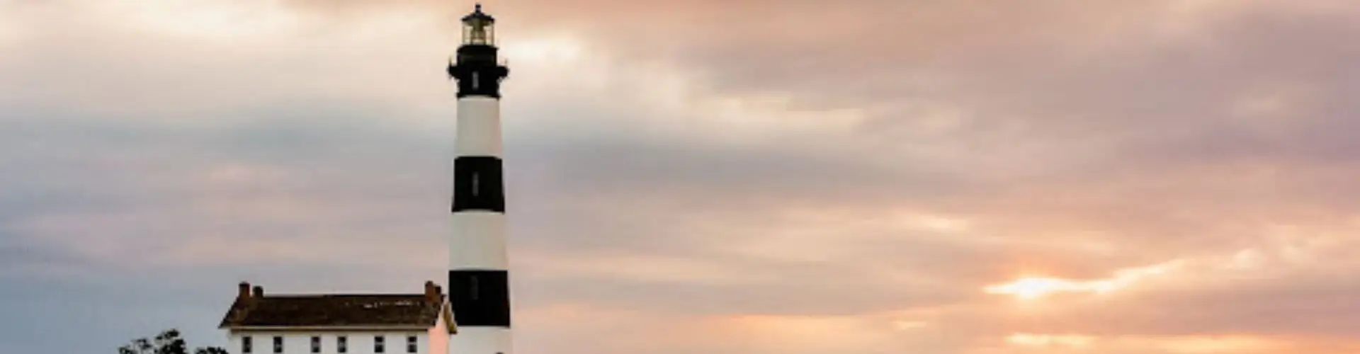 A black and white banded lighthouse and its keeper's house silhouetted against a dusky, cloud-streaked sky