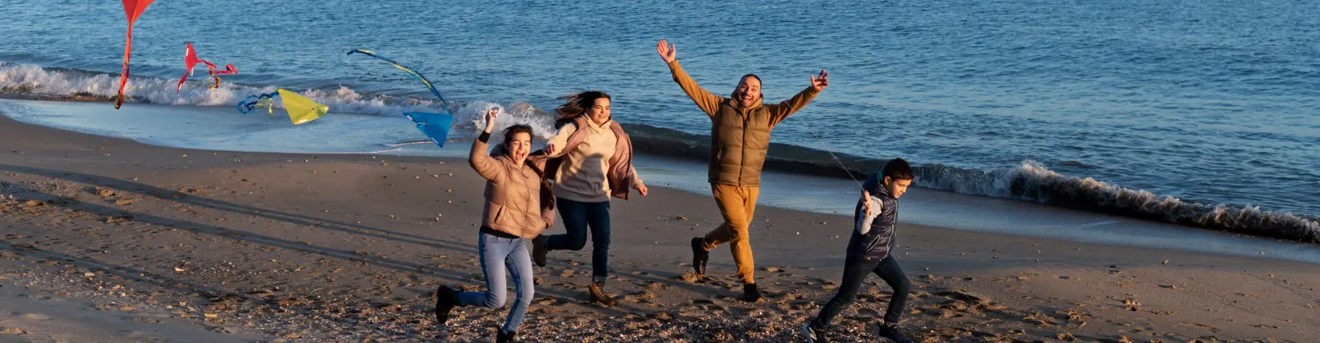 A family running together along the shoreline, kites flying above the dunes behind them