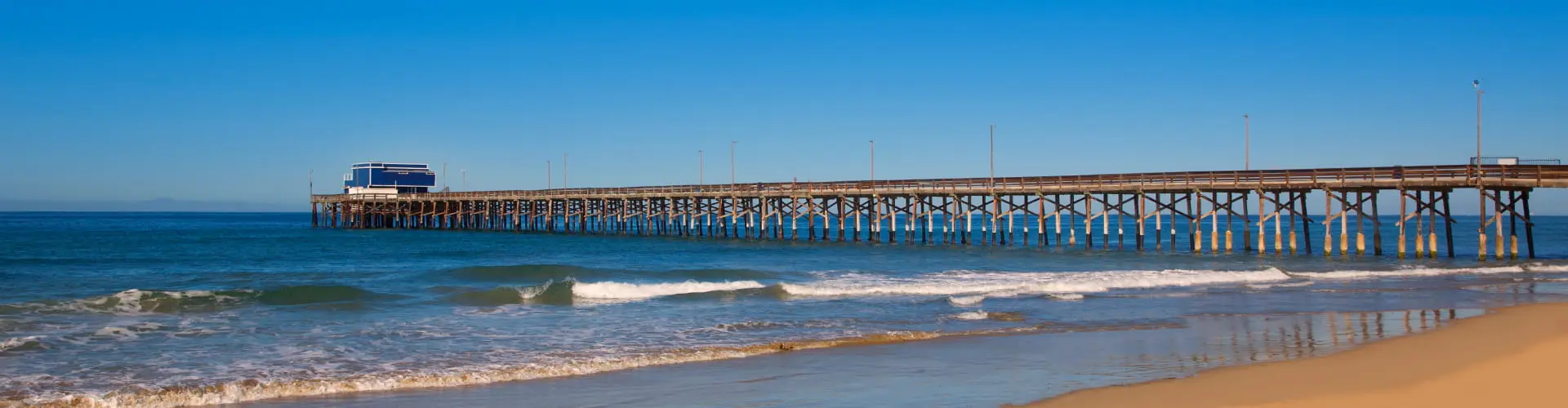 A long wooden fishing pier reaching out over blue ocean from an empty sunlit beach