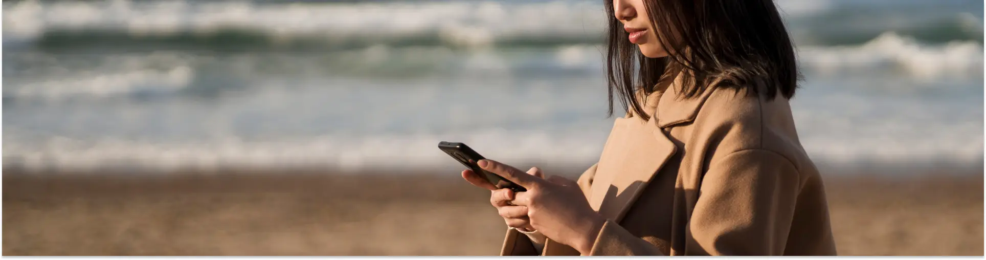 A woman in a camel coat reading her phone on a windy beach, waves breaking behind her