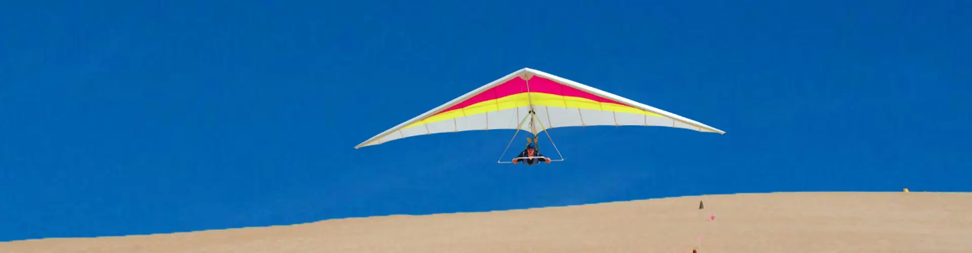 A red and yellow hang glider soaring over a sand dune against a cloudless blue sky