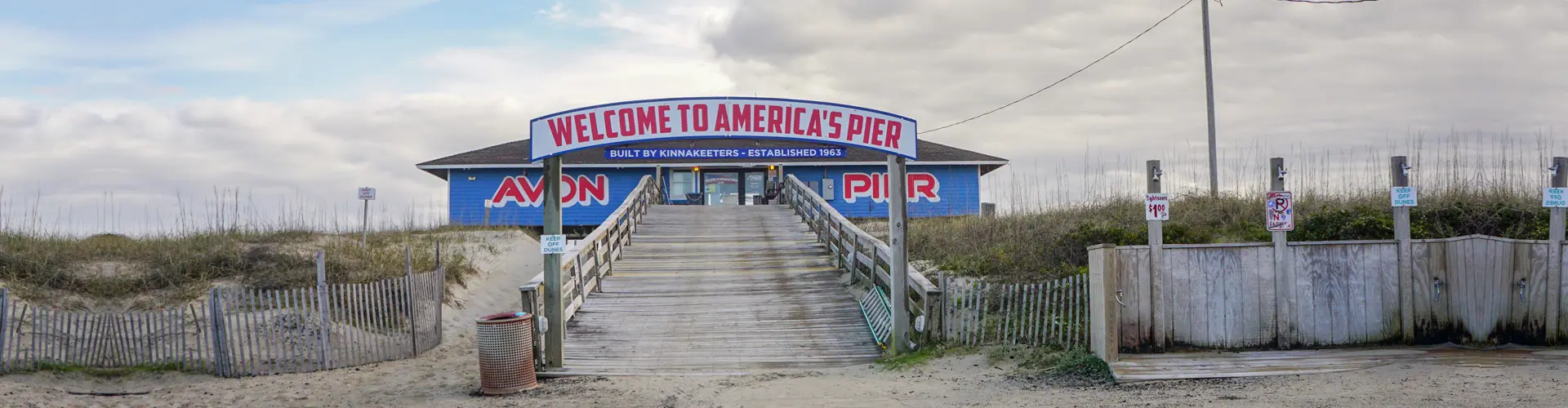 The boardwalk through the dunes to Avon Pier, its blue pier house under an arched Welcome to America's Pier sign