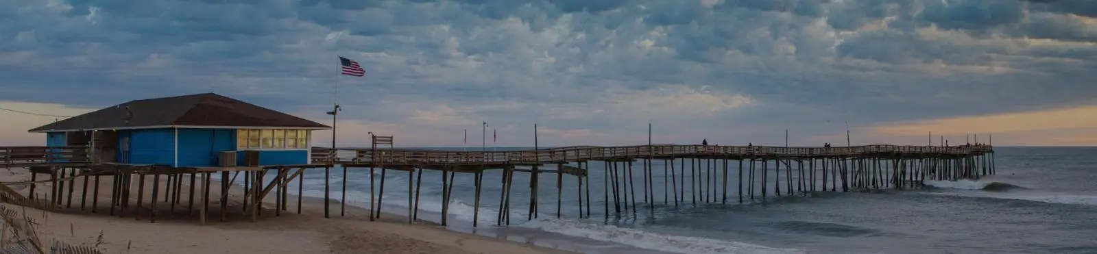 A long fishing pier with a blue pier house and a flag, running out over a grey, choppy ocean