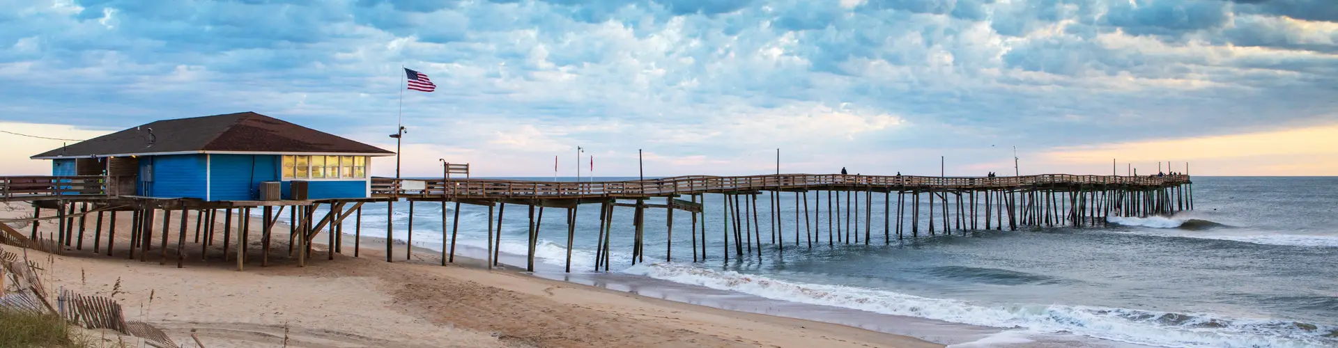 A blue-roofed pier house on pilings and its pier stretching into the surf, seen from the beach under cloud
