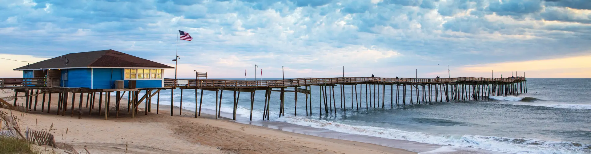 A blue-roofed pier house on pilings and its pier stretching into the surf, seen from the beach under cloud