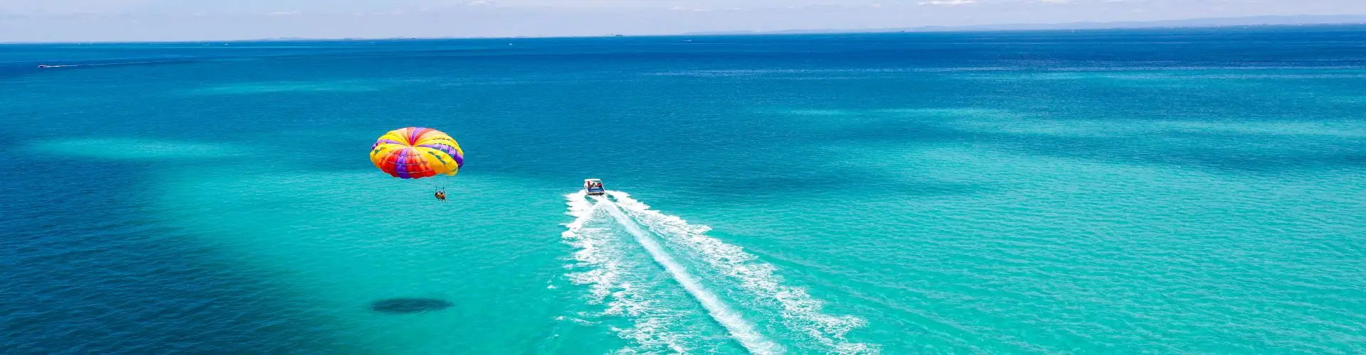 A parasail seen from above over brilliant turquoise water, the tow boat trailing a long white wake