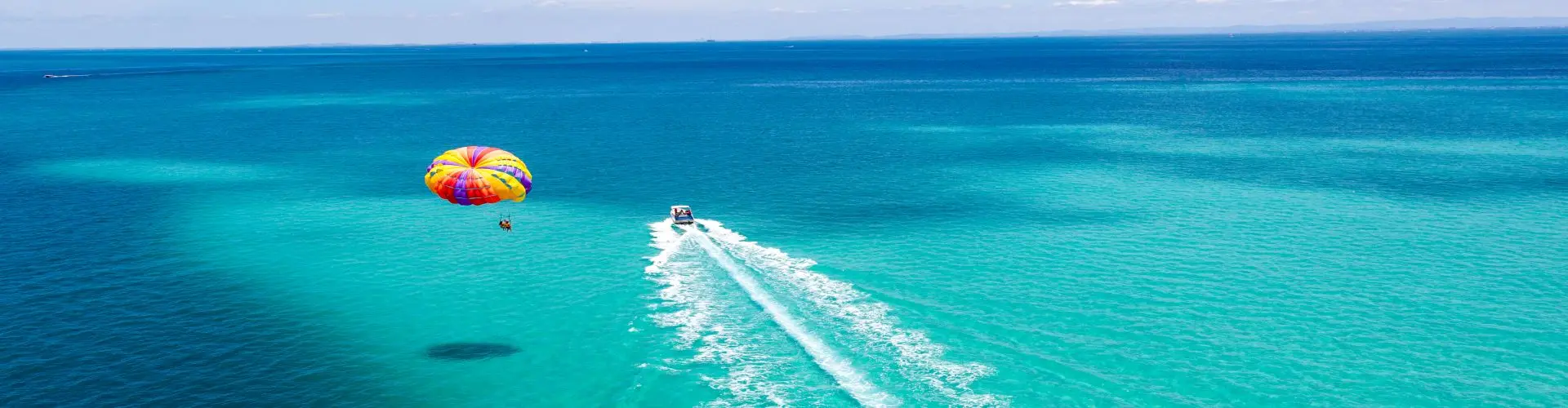 A parasail seen from above over brilliant turquoise water, the tow boat trailing a long white wake