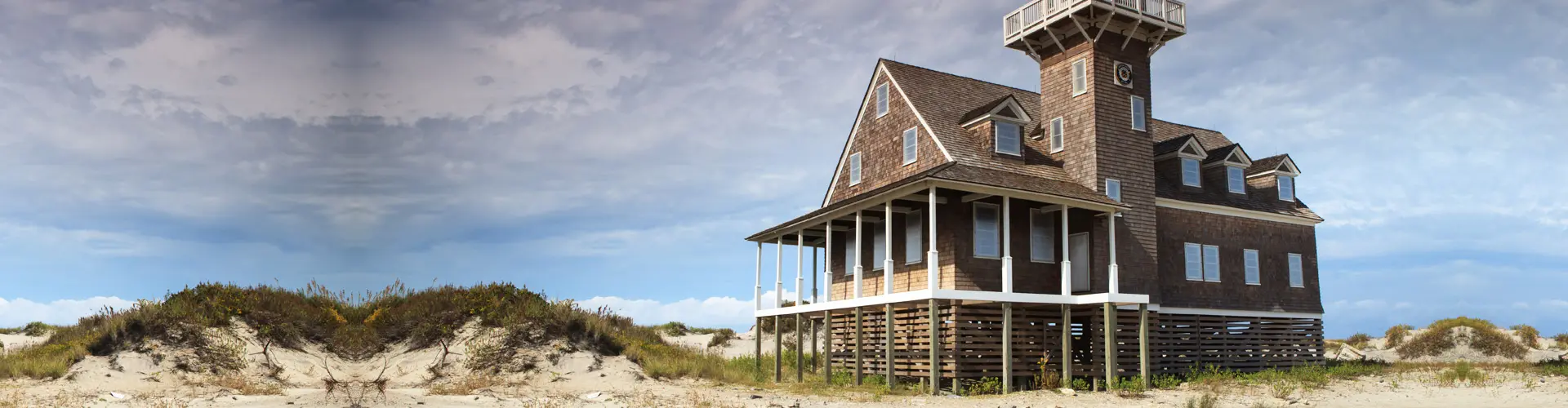 A cedar-shingled life-saving station on the dunes, its rooftop lookout tower against a big cloudy sky