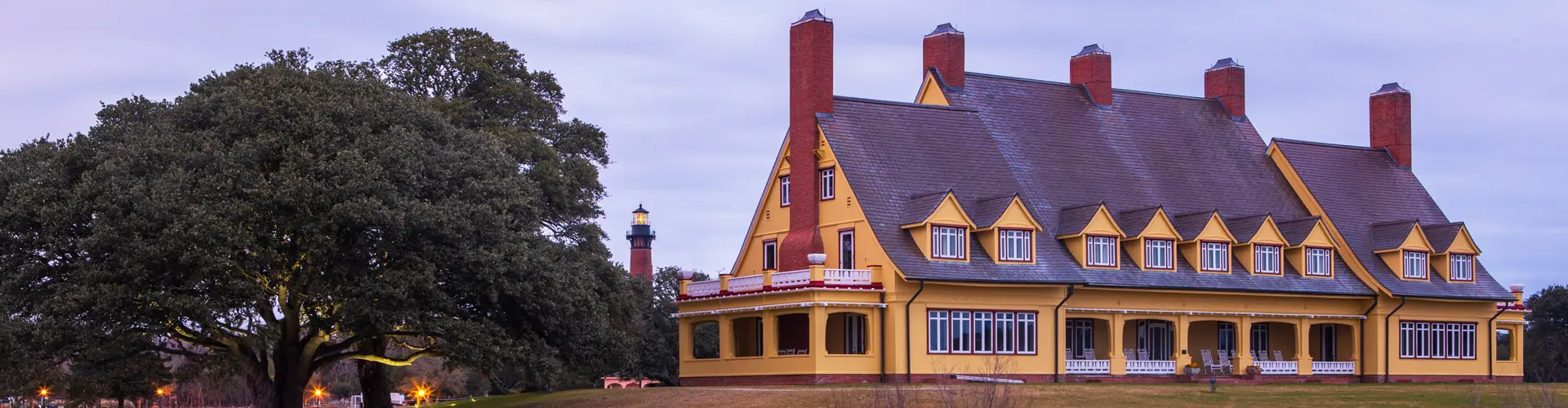 A yellow gabled mansion with tall red-brick chimneys at dusk, the Currituck Beach Lighthouse lit behind the live oaks