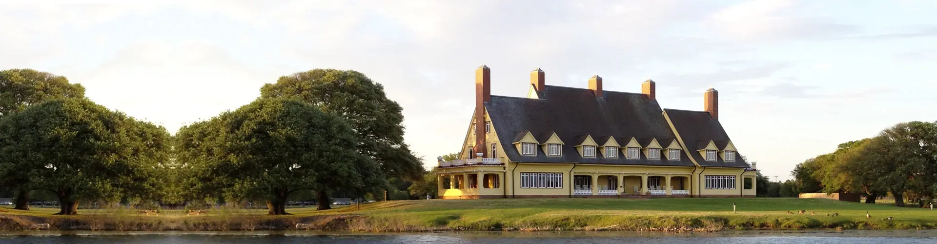 A long yellow mansion with a steep slate roof and brick chimneys across water and lawn, live oaks alongside in evening light