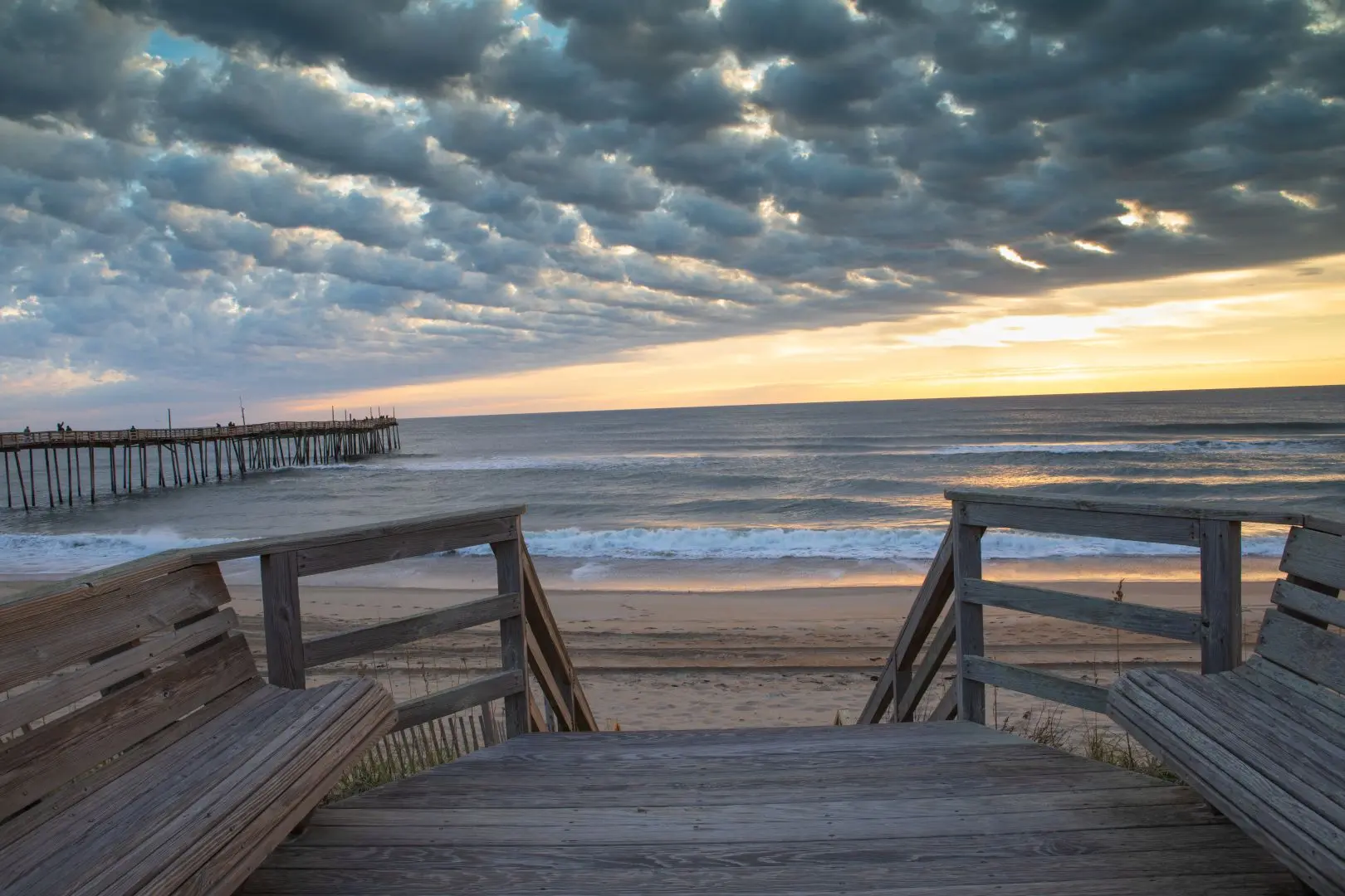 Weathered boardwalk steps down onto the beach at sunrise, a pier in the distance under dramatic cloud