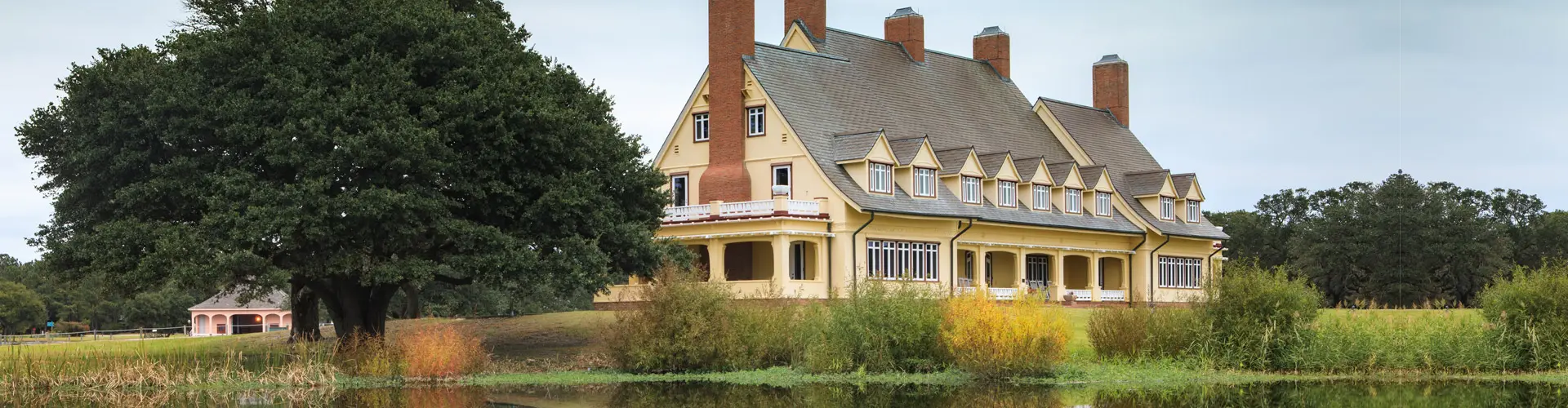 A yellow gambrel-roofed mansion with tall brick chimneys behind a reedy pond and a spreading live oak