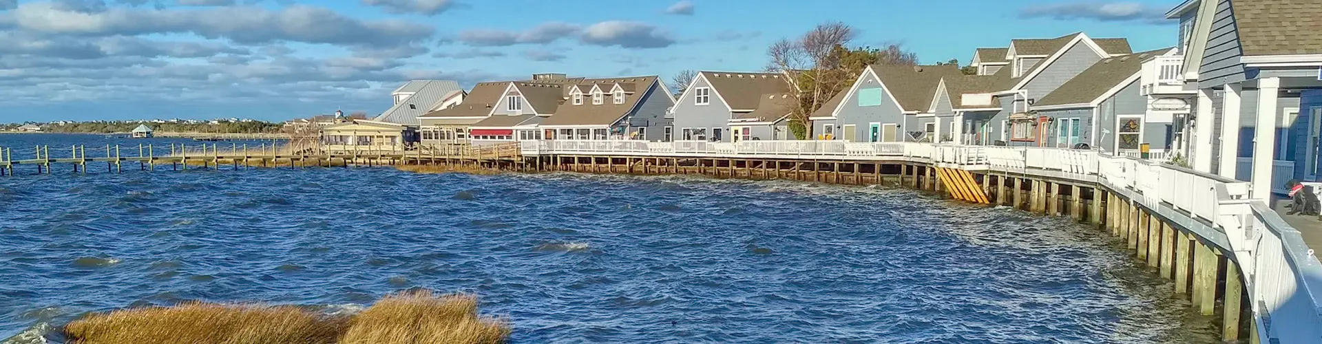 Grey-shingled shops and restaurants along a soundfront boardwalk, wind-chopped blue water in the foreground