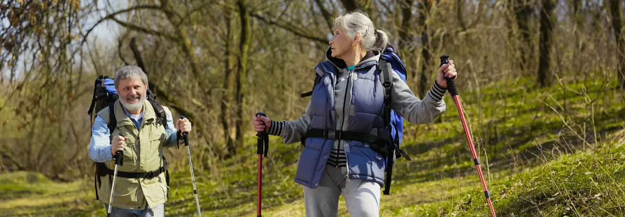 An older couple with rucksacks and walking poles striding along a green woodland trail