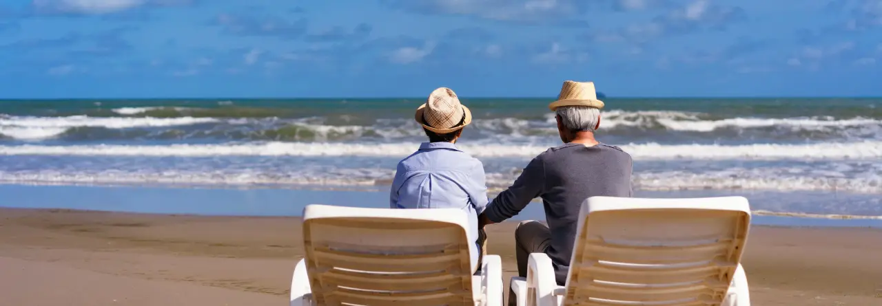 Two older men in sun hats sitting in white loungers on the sand, watching a blue sea