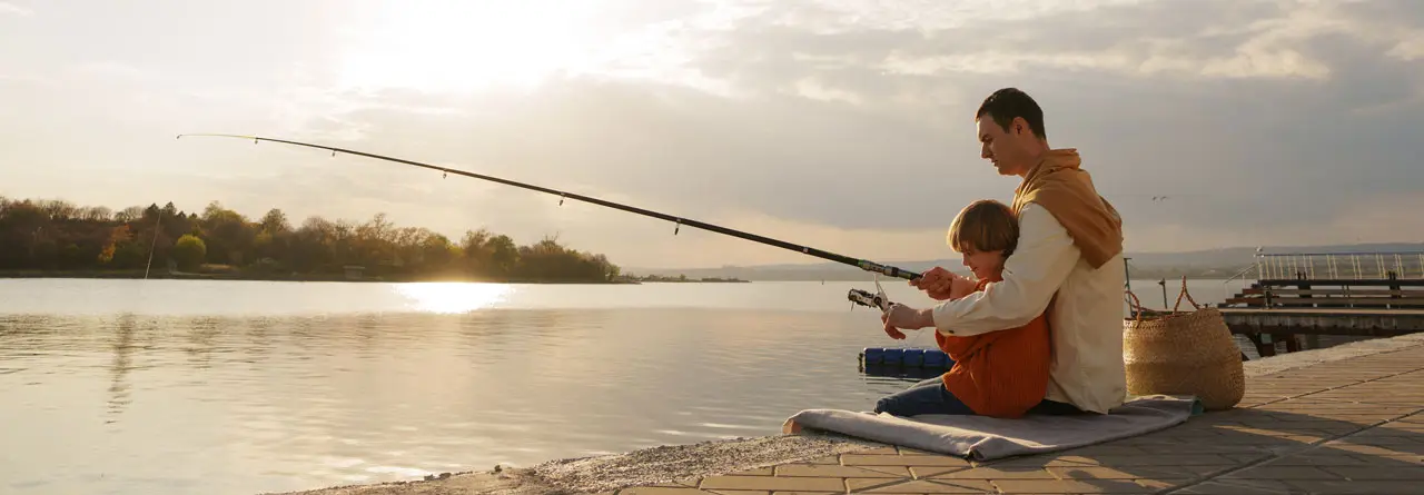 A man and a small child fishing from a wooden dock over flat golden water at sunrise
