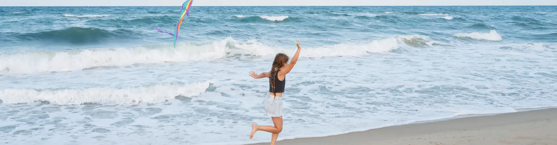 A girl running barefoot through the shallows, flying a rainbow kite above the breaking waves