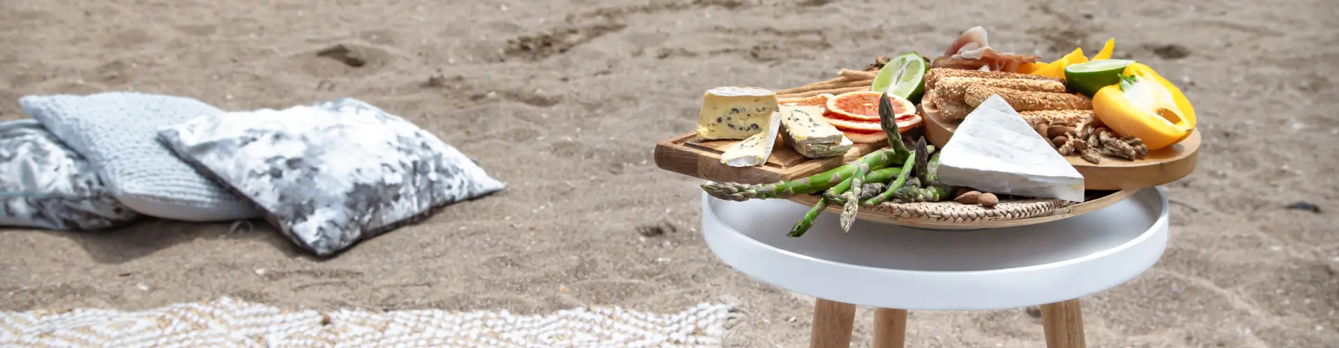 A round wooden board of cheese, bread and fruit on a low table set up on the sand