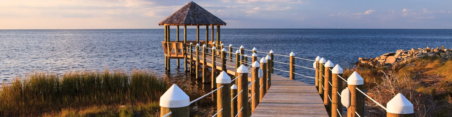 A wooden walkway with white-capped posts running out to a shingled gazebo over calm sound water at golden hour