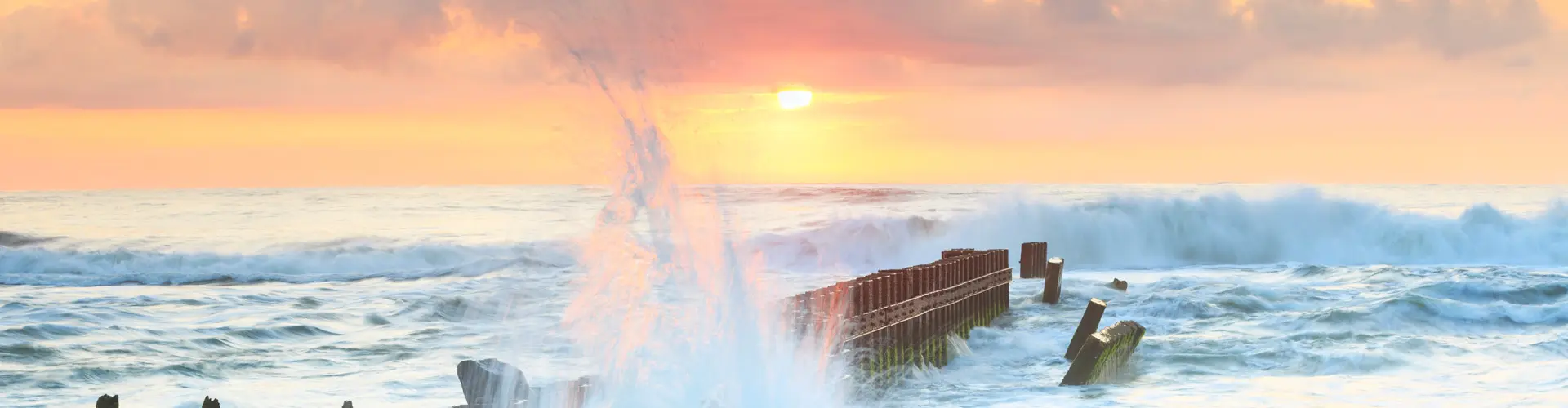 A wave bursting over the rusted iron remains of a wreck in the surf as the sun rises in a pink sky