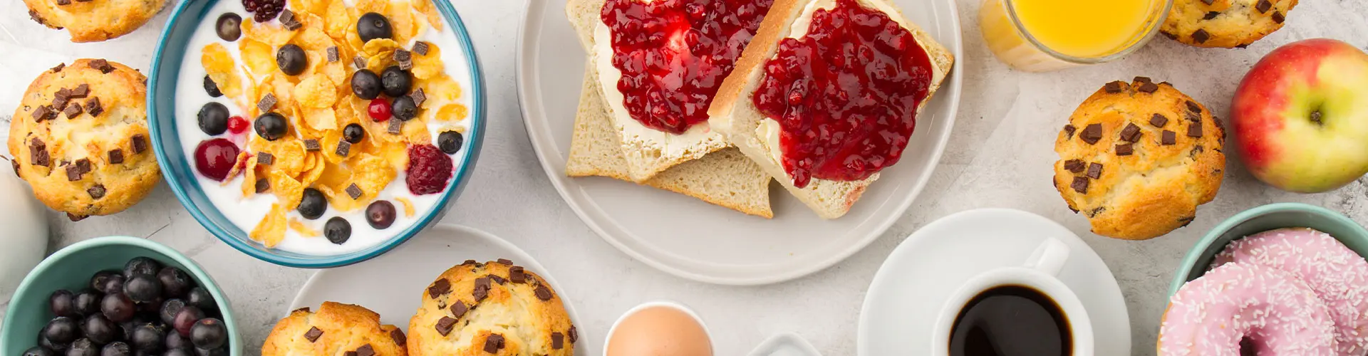An overhead spread of muffins, berries, cheesecake slices, doughnuts, coffee and orange juice