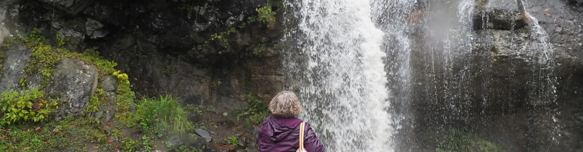 A walker in a purple jacket standing below a tall waterfall spilling down mossy rock