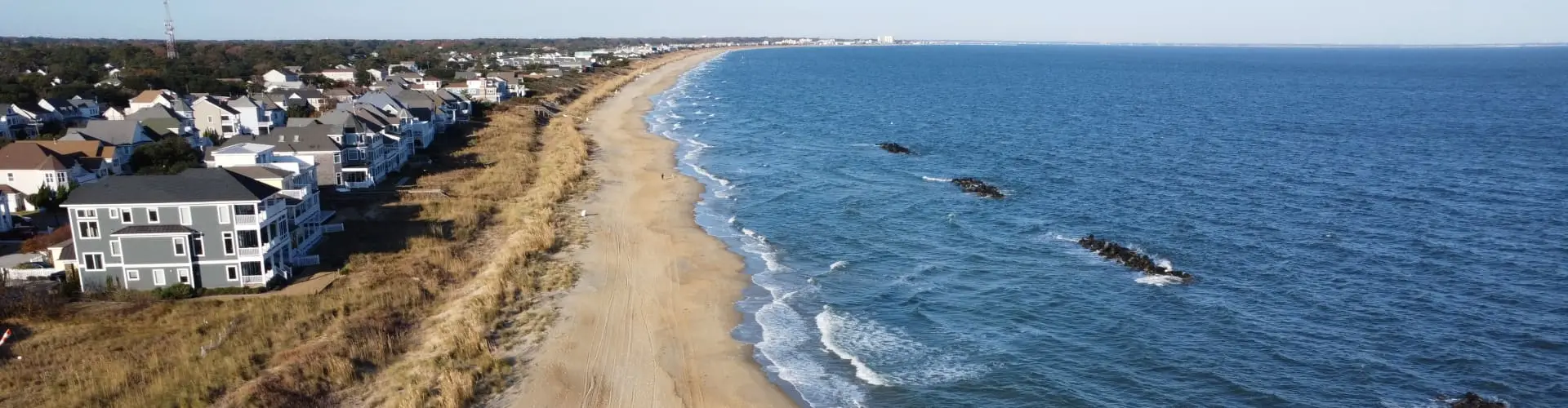 Beachfront houses ranged behind the dune line above a long strip of sand and blue ocean, seen from the air