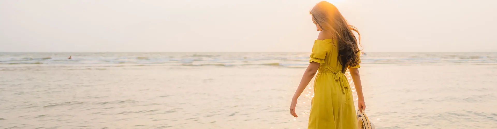 A woman in a yellow sundress walking at the water's edge in soft hazy light, hair lifting in the breeze