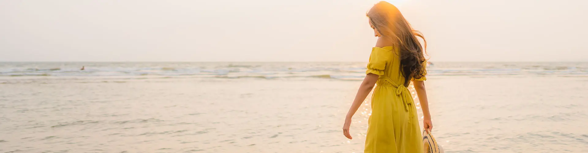 A woman in a yellow sundress walking at the water's edge in soft hazy light, hair lifting in the breeze