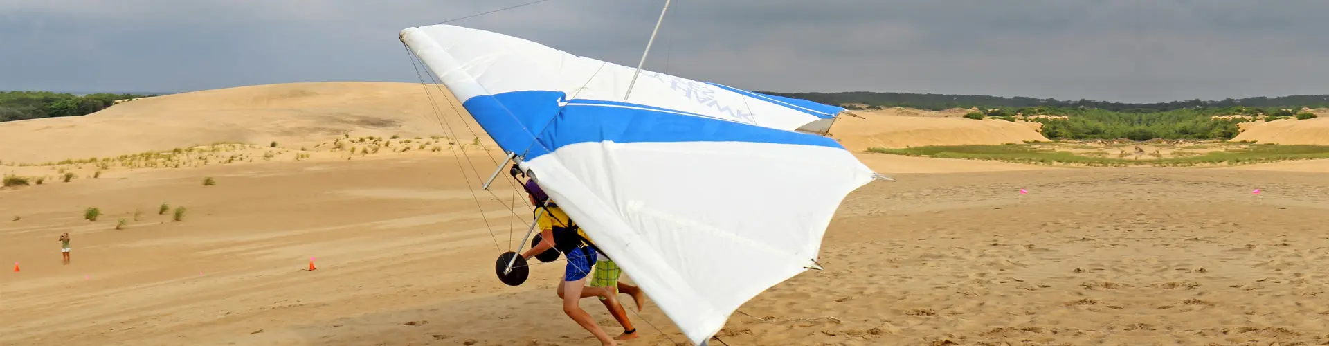 A blue and white hang glider lifting off a bare sand dune, its pilot harnessed beneath the wing