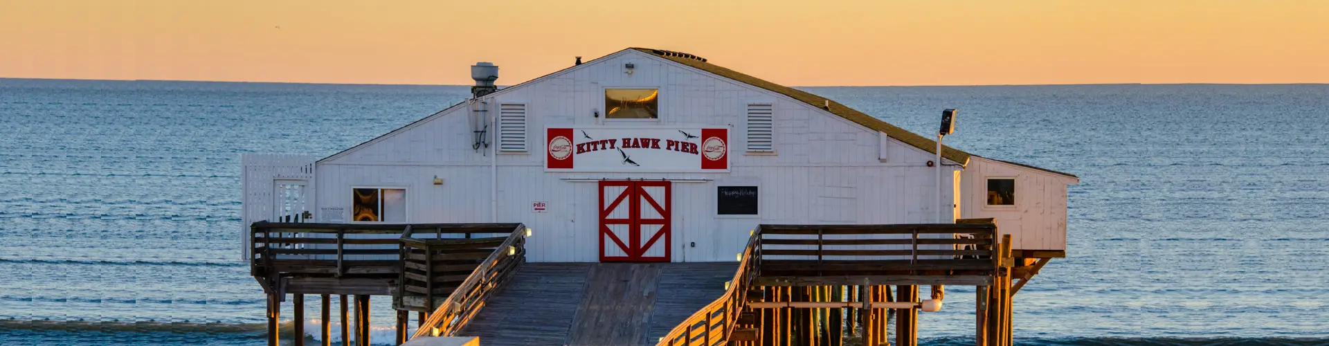 The white clapboard Kitty Hawk Pier house with red barn doors, standing on pilings over a calm sea at sunset