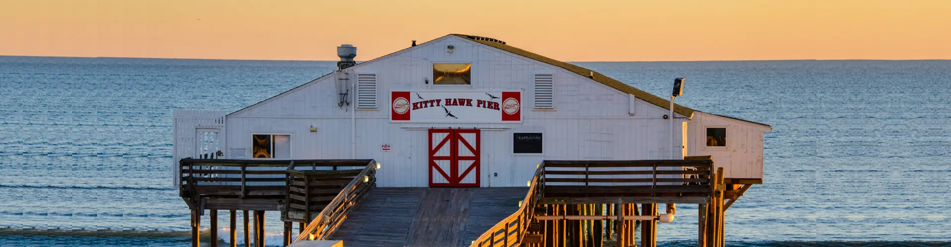 The white clapboard Kitty Hawk Pier house with red barn doors, standing on pilings over a calm sea at sunset
