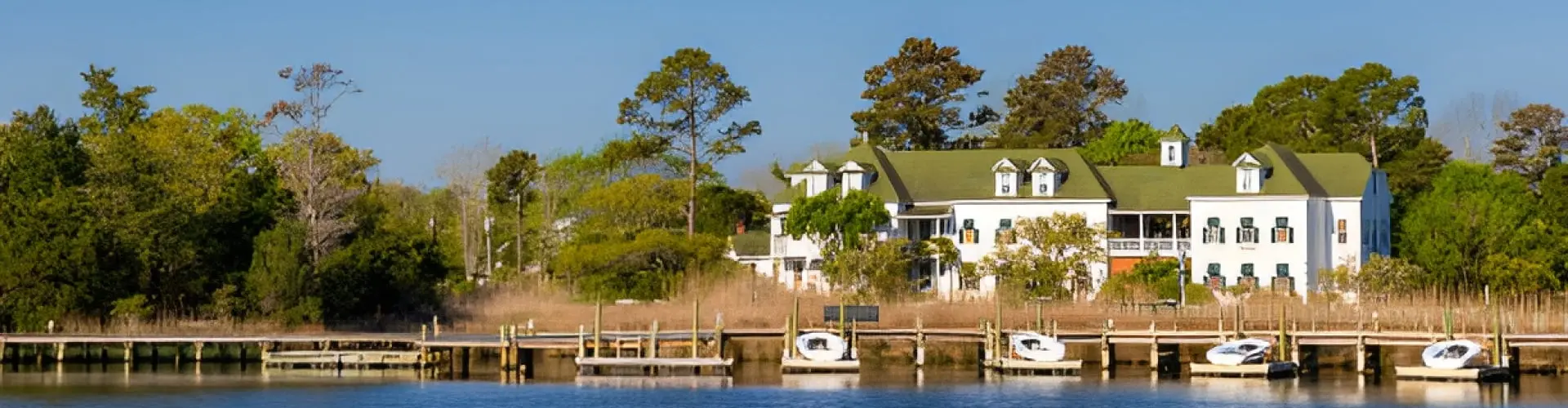 A long white green-roofed waterfront building among trees, small white sailboats moored at wooden docks in front