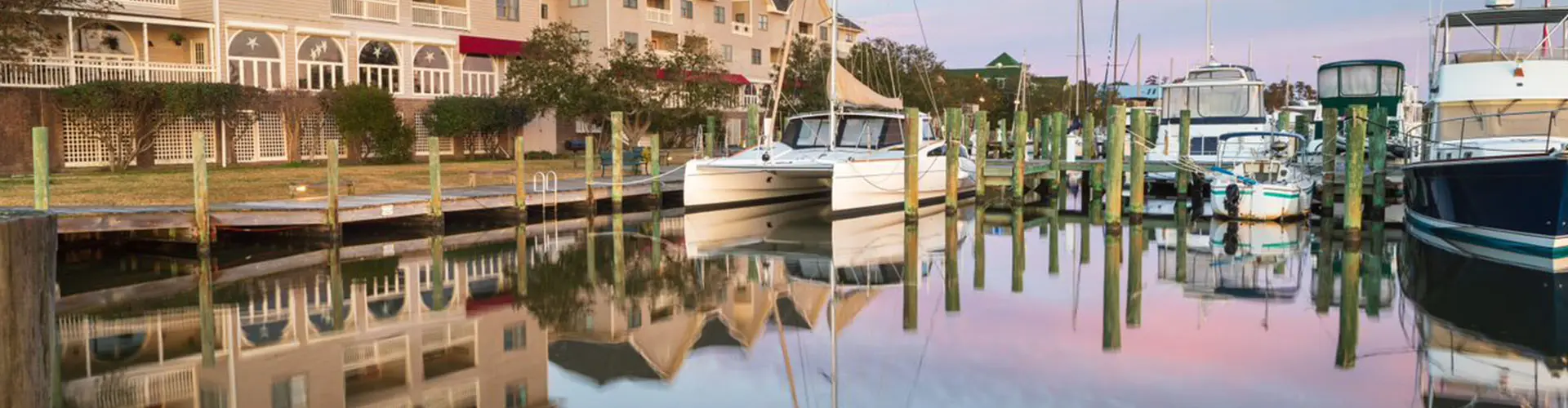 Boats moored along a wooden dock in a quiet marina, waterfront buildings mirrored in still pastel water