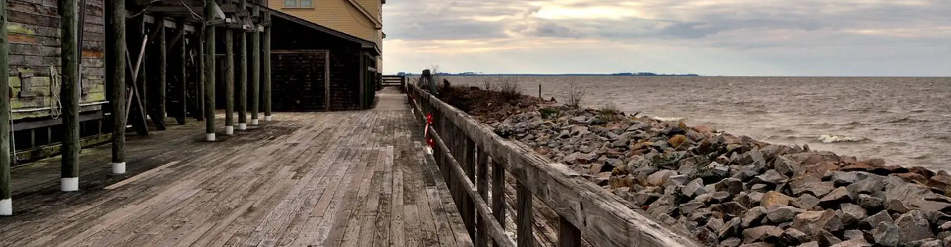 A weathered boardwalk beside a rock revetment along a grey, wind-ruffled sound, old pilings and a yellow clapboard building on stilts