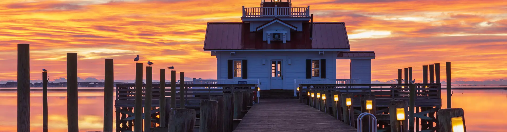 The white cottage-style Roanoke Marshes Lighthouse at the end of a lamp-lit boardwalk under a blazing orange sunrise