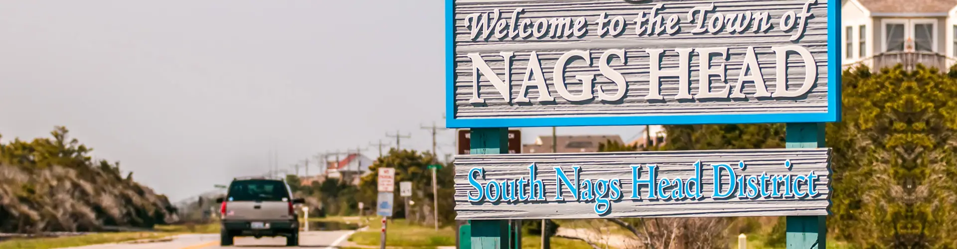 A roadside sign welcoming drivers to the town of Nags Head, South Nags Head District, a pickup truck passing behind