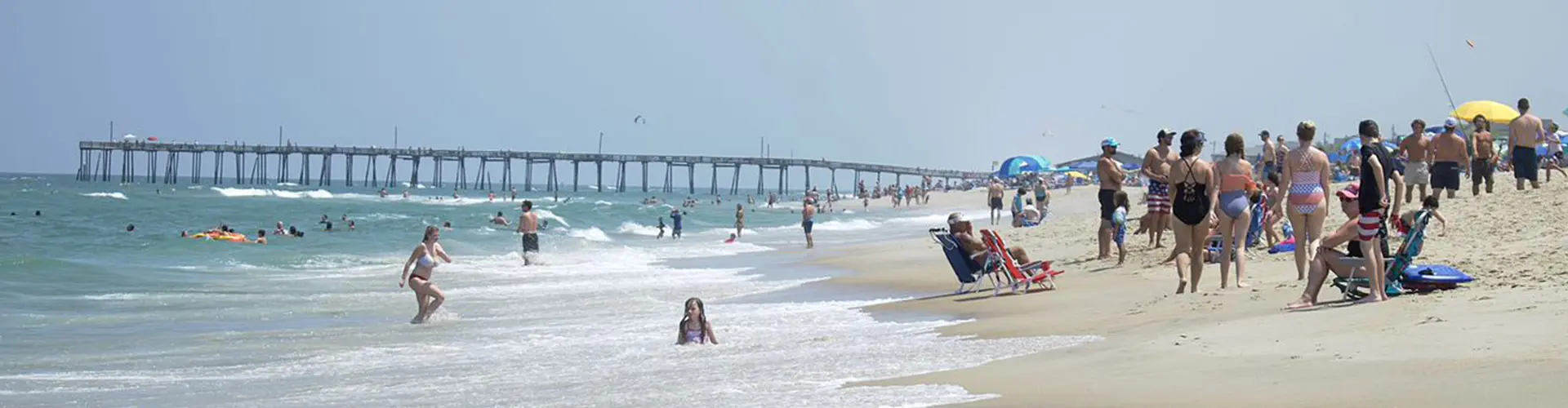 A busy summer beach of swimmers, umbrellas and towels, a fishing pier stretching out in the distance