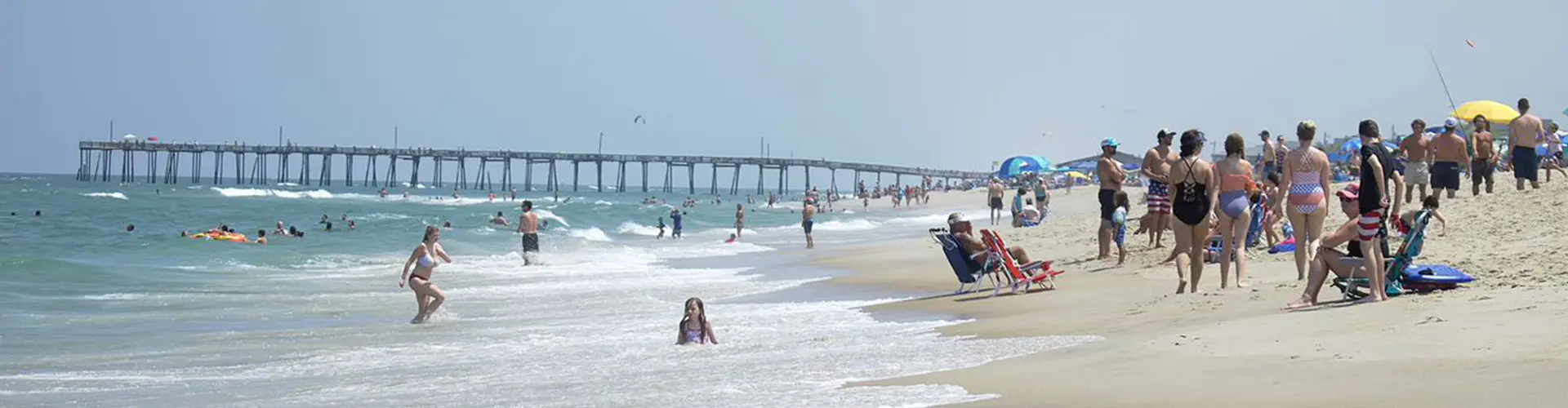 A busy summer beach of swimmers, umbrellas and towels, a fishing pier stretching out in the distance