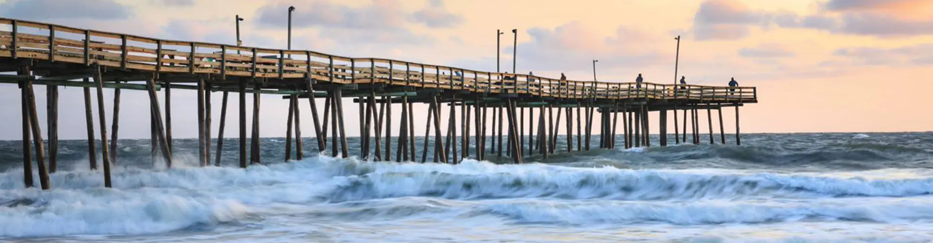 Waves rolling under a long fishing pier beneath a soft pink and gold dawn sky
