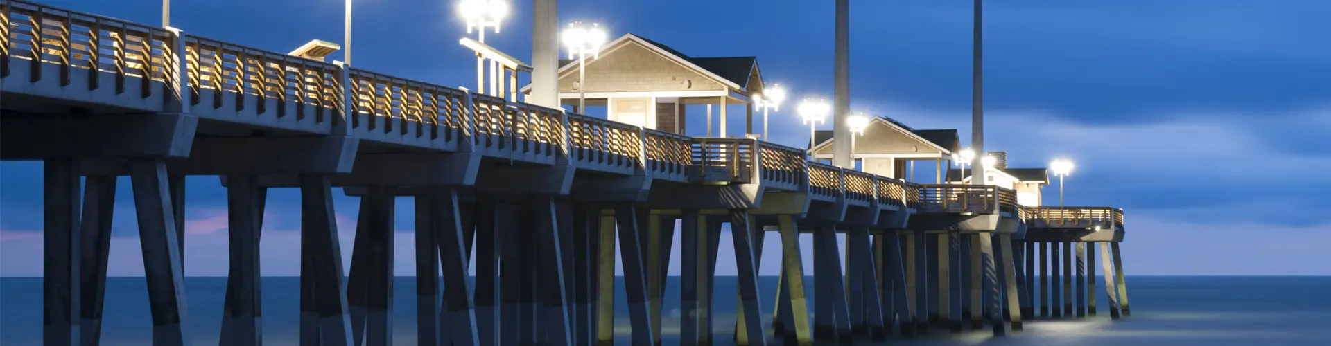 A fishing pier lit up at blue hour, its lamps glowing above the pilings and dark surf