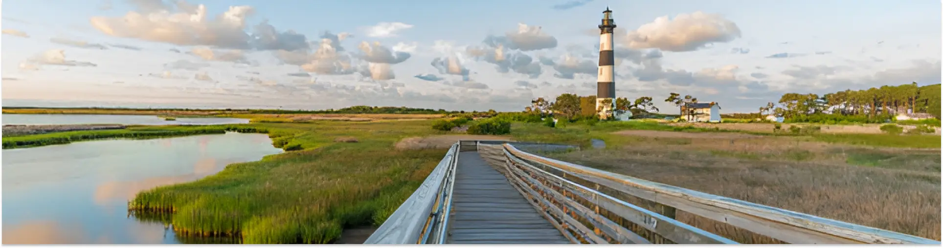 A boardwalk crossing green marsh and still water towards the black-and-white banded Bodie Island Lighthouse