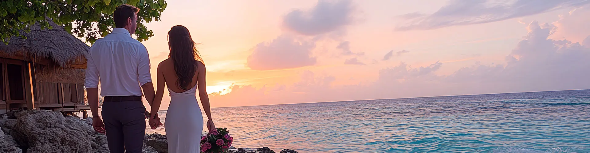A couple holding hands beneath a thatched shelter, watching a pink and gold sunset over the sea
