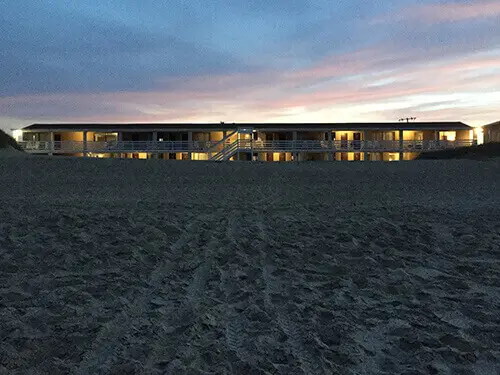 Outer Banks Motor Lodge at dusk seen from the beach, its windows lit above the sand
